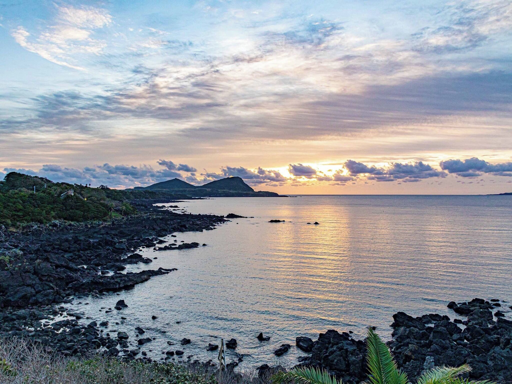 Un paisaje costero con costa rocosa, mar tranquilo y montañas distantes bajo un cielo parcialmente nublado.
