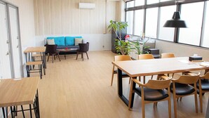Un espacio de oficina moderno con suelo de madera, un sofá con cojines azules, una mesa de comedor con sillas y grandes ventanales que dejan entrar luz natural.
