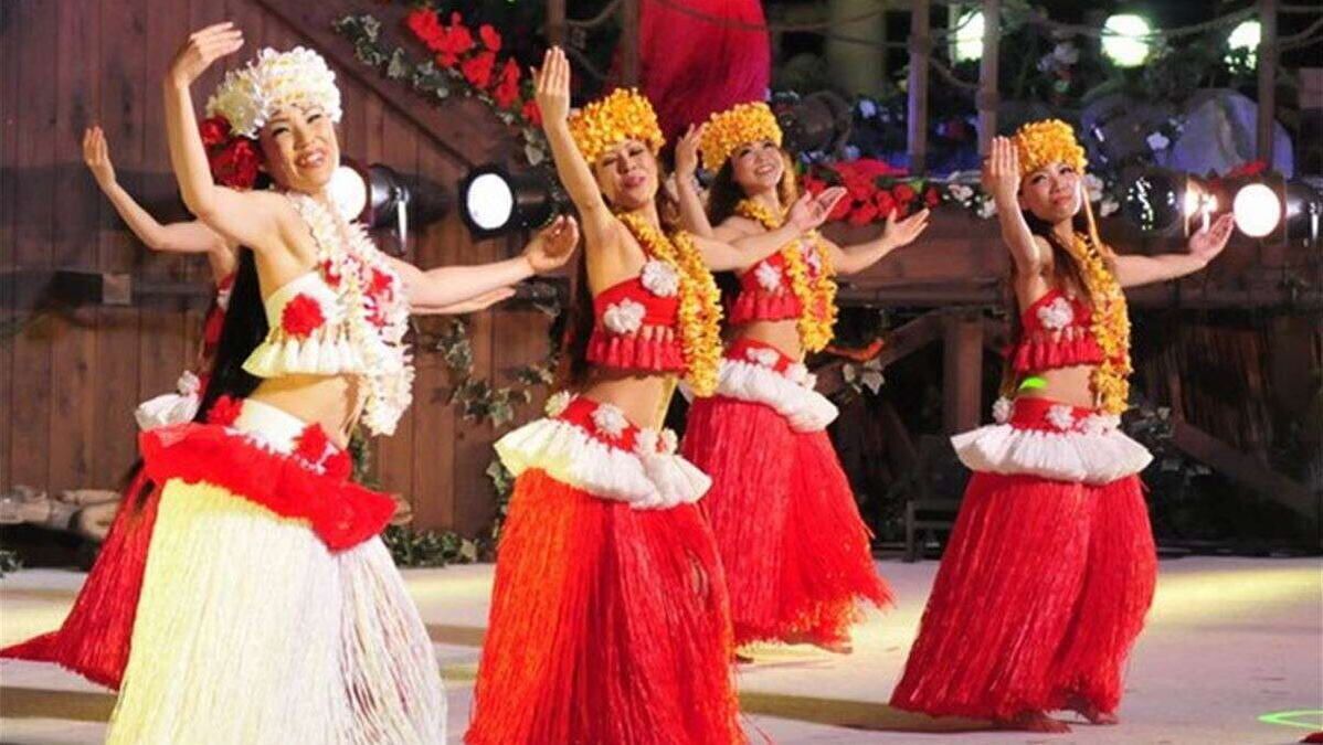 A group of performers in traditional costumes with floral garlands, performing on a stage with a wooden backdrop and stage lights.