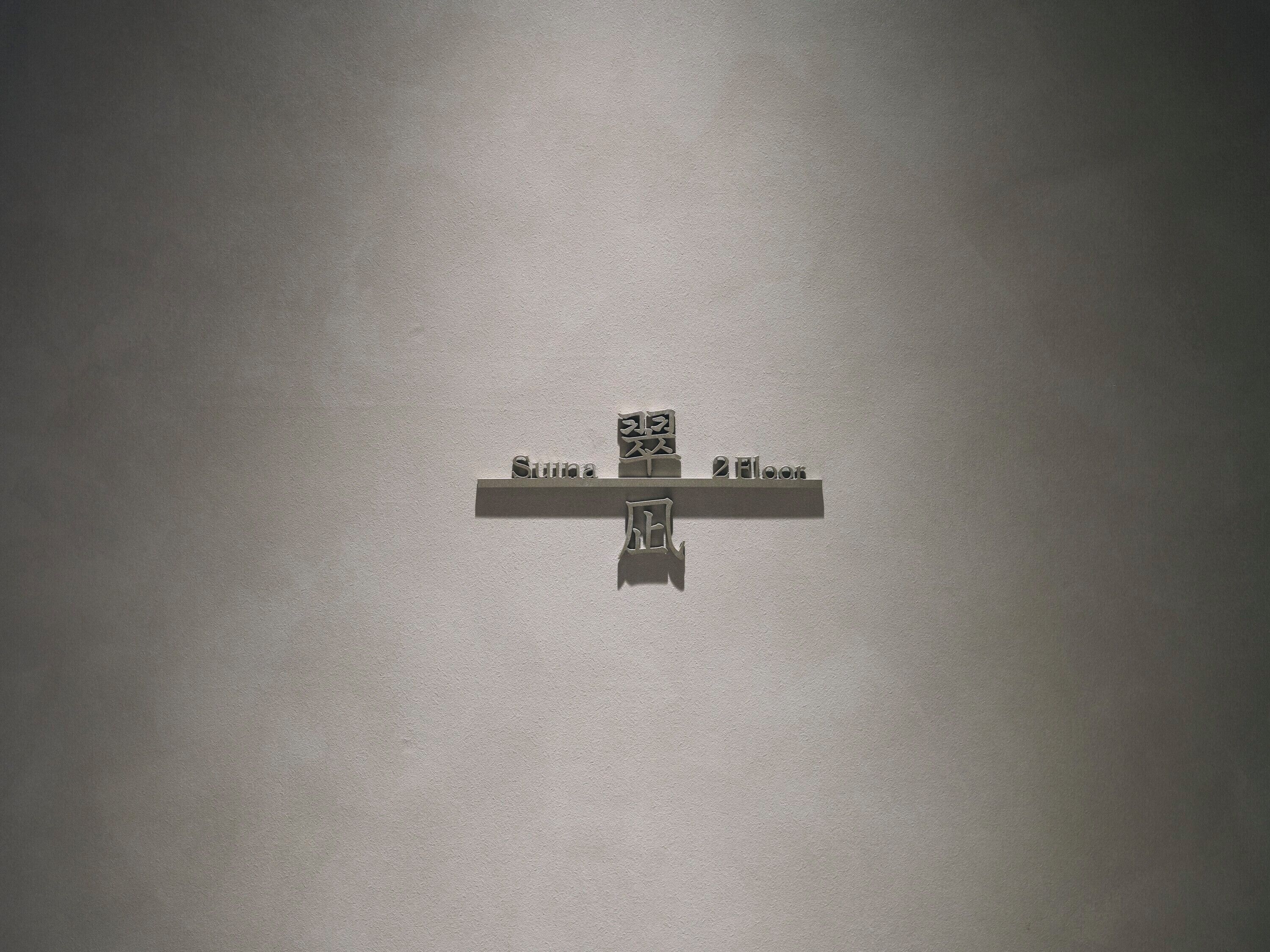 A minimalist cross-shaped sign with the words 'Suna' and '2nd floor' on a plain wall.