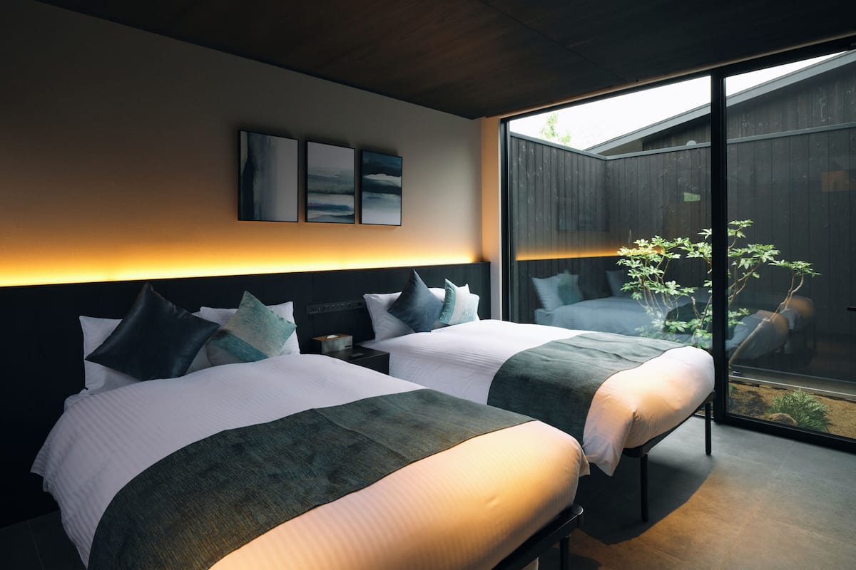 A modern hotel room with two beds, a large window, and a view of the outdoors.
