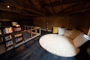 Um quarto acolhedor com teto de madeira, um tapete redondo, uma cama com almofadas e uma estante repleta de livros.