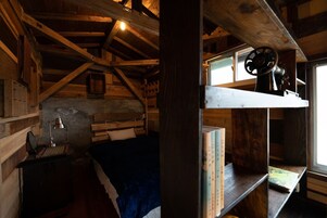 Interior rústico de cabana de madeira com cama no mezanino, escrivaninha com abajur e prateleira com livros.