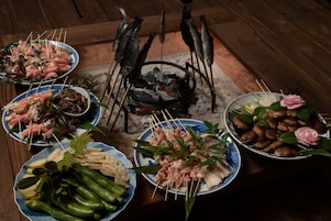 Disposição tradicional de refeição japonesa com diversos pratos e espetos de peixe e carne, dispostos ao redor de uma churrasqueira a carvão.