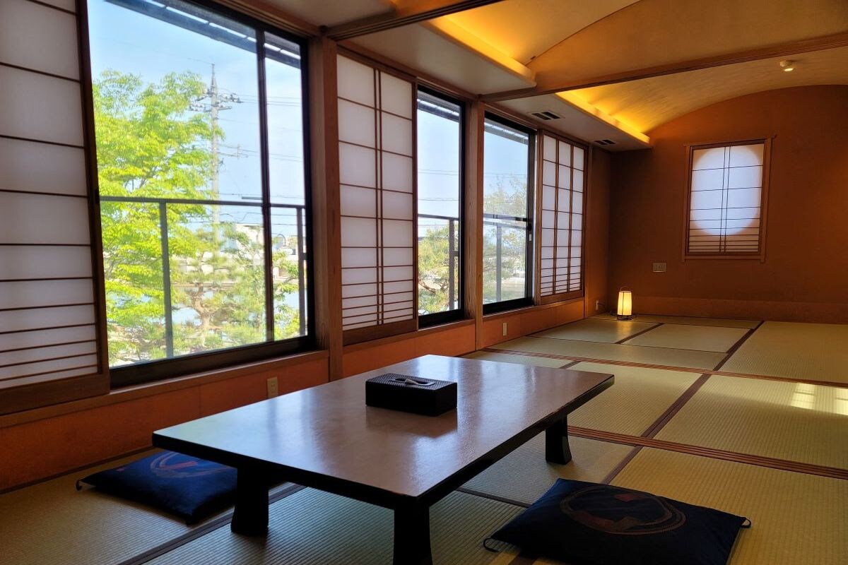 A tatami mat room with a low table and cushions, large windows with shoji screens, and a view of greenery outside.