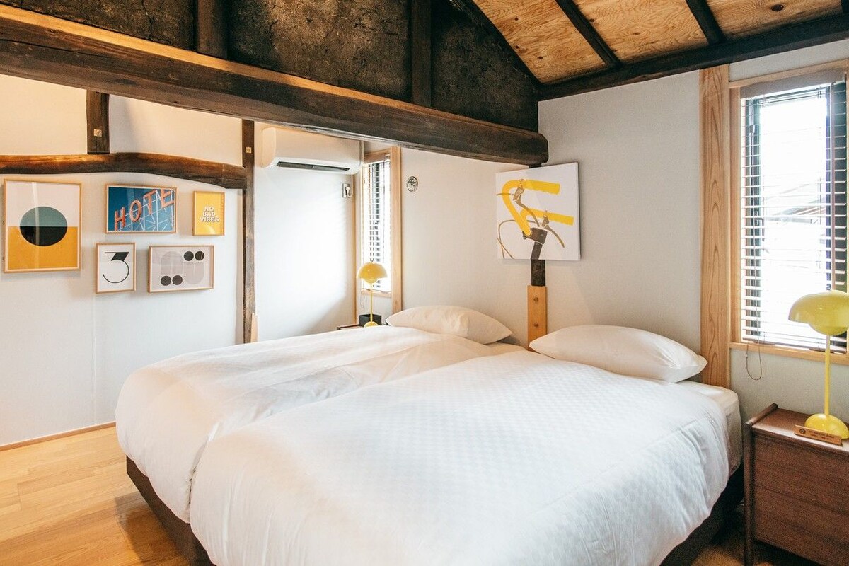 A bedroom with a large bed, wooden beams, a window with blinds, and wall art.