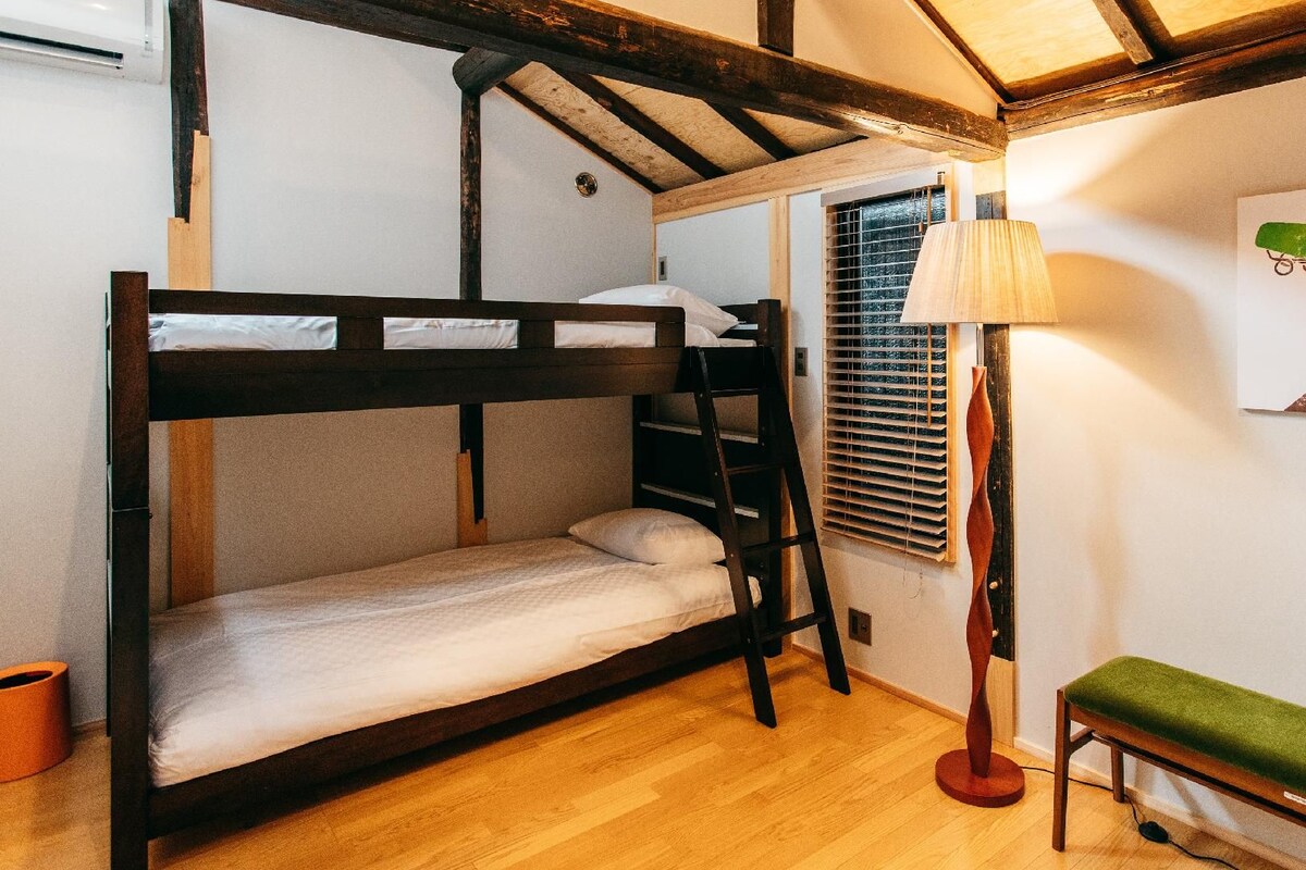 A bunk bed room with a wooden ladder, a bench, a floor lamp, and a window with blinds.