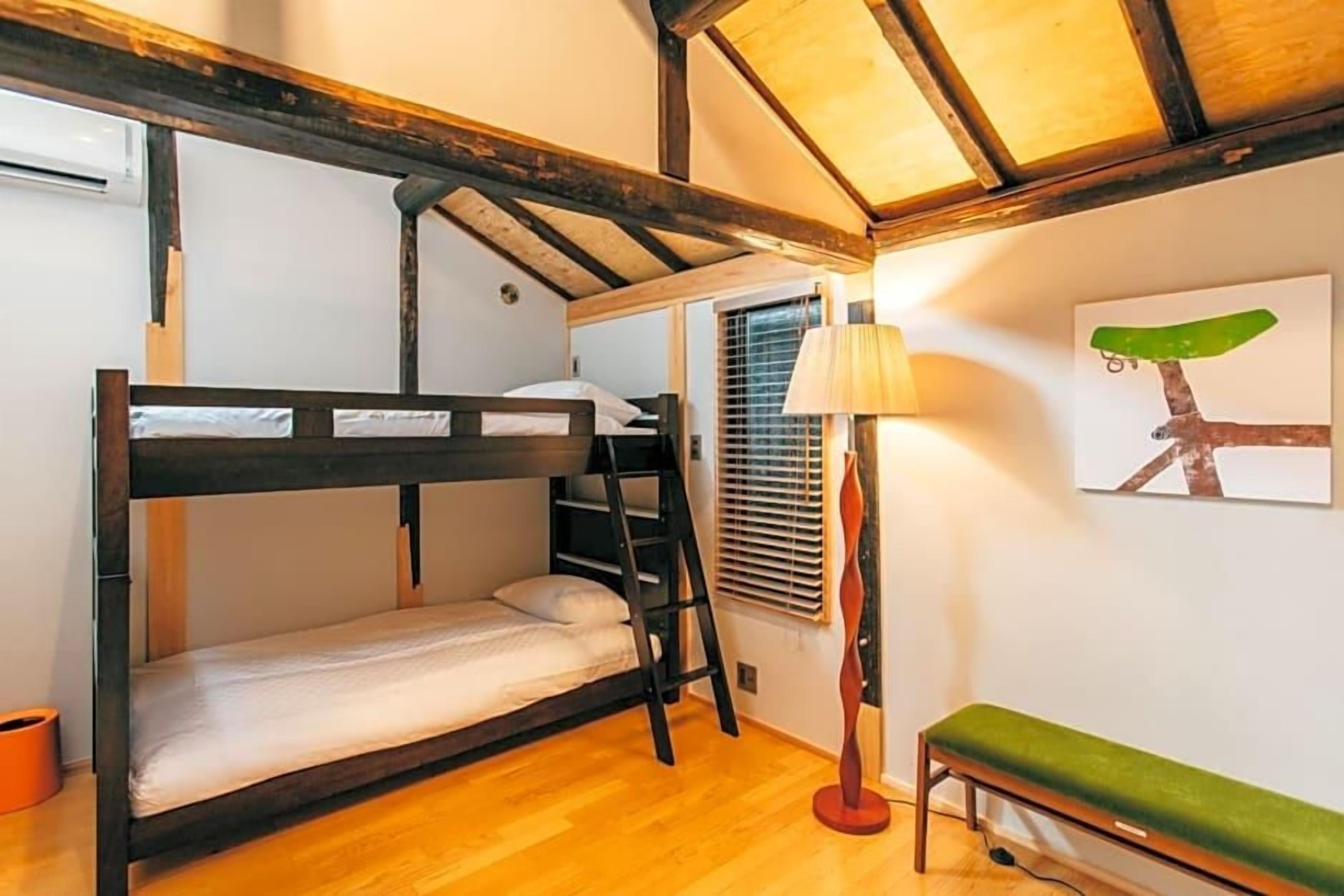 A bunk bed room with a wooden floor, a ladder, a bench, a lamp, and a wall art piece.