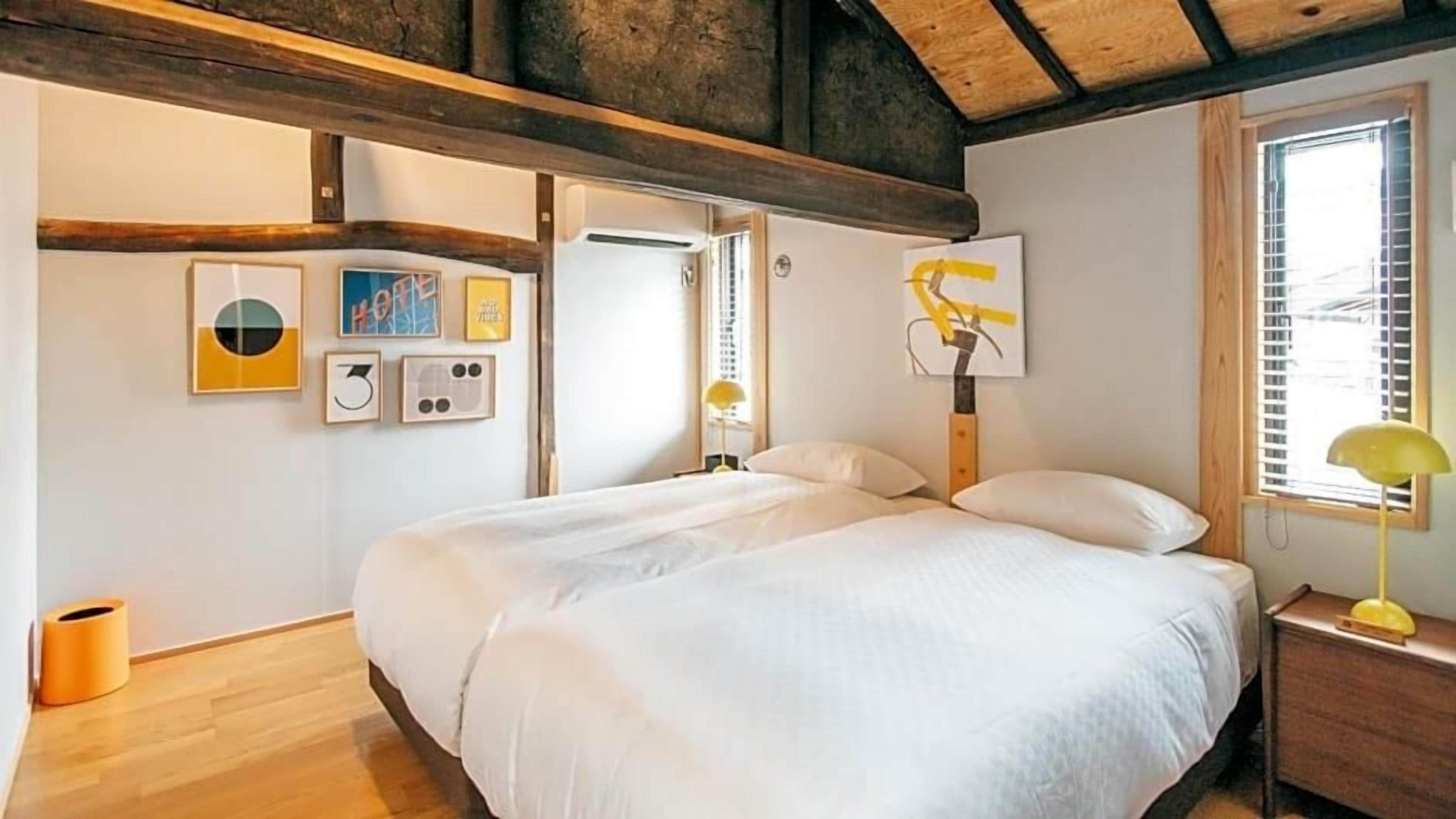 A bedroom with a large bed, wooden beams, a window with blinds, and wall-mounted artwork.