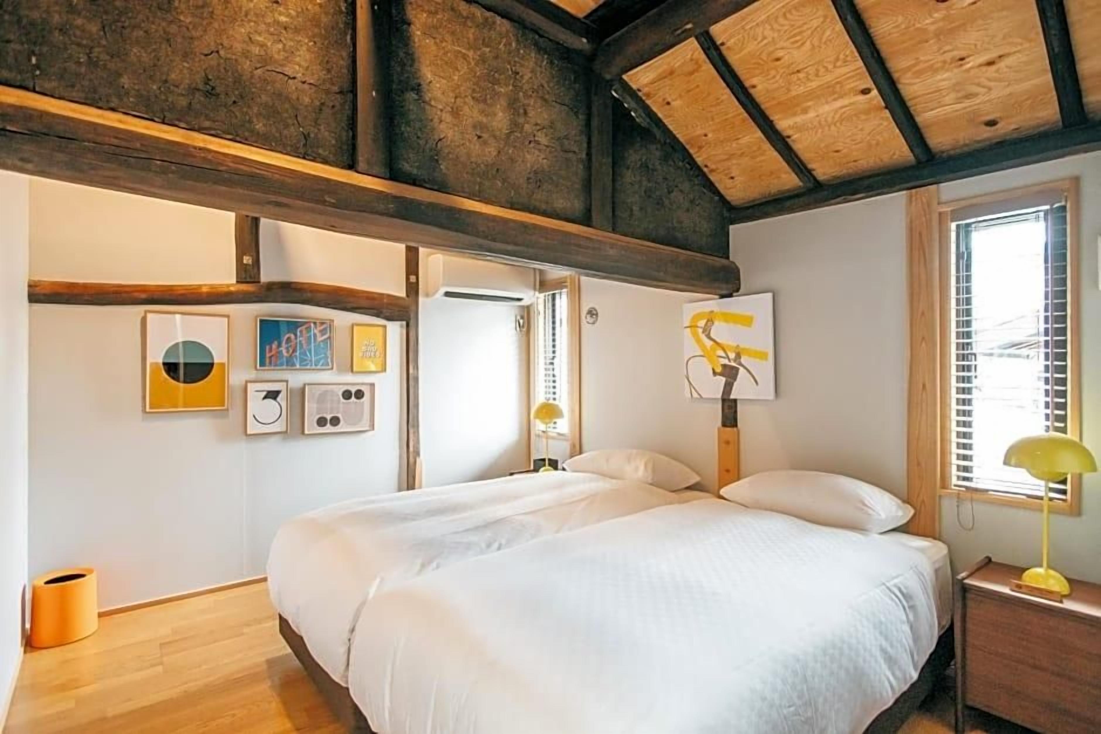 A bedroom with a large bed, wooden beams, a window with blinds, and wall art.
