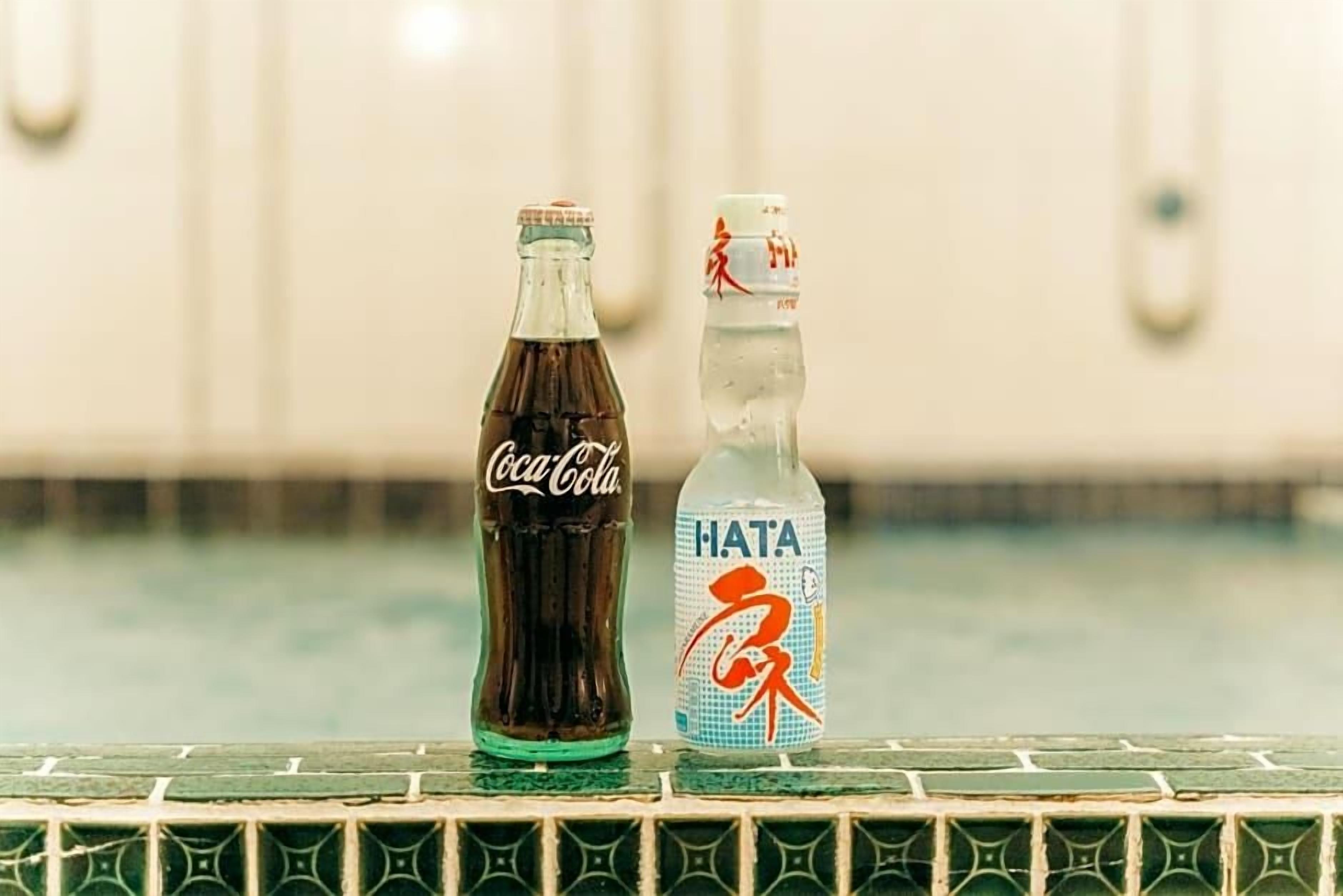 Two bottles, one labeled Coca-Cola and the other Hata, placed on a tiled surface.