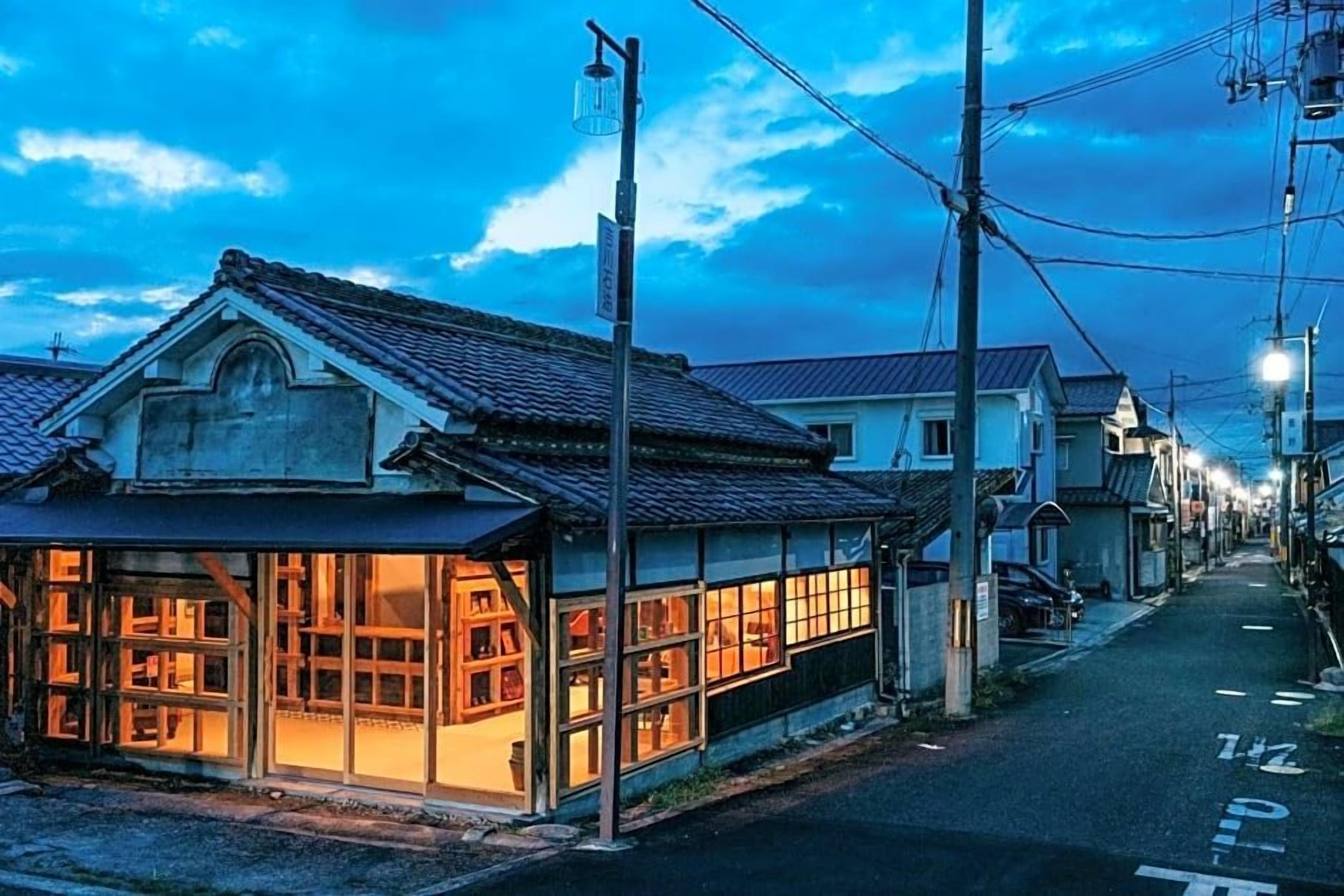 A traditional Japanese building with large windows and a tiled roof, located on a narrow street with other residential buildings.