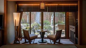 A room with a traditional Japanese-style interior, featuring a tatami mat floor, a low table with two chairs, a large window with a view of a garden, and a hanging lamp. - Tokisumika (Komagane)