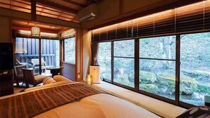 A bedroom with a large bed, a desk, and a chair, with a view of a forest through large windows. - Tokisumika (Komagane)