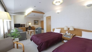 Quarto de hotel com duas camas, uma escrivaninha, uma cadeira, uma televisão e uma planta.