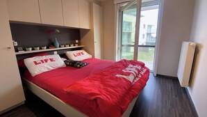 A bedroom with a bed, a shelf with small plants, and a window with a view of buildings. - Apartment with sunny terrace in Oostduinkerke (OOSTDUINKERKE)