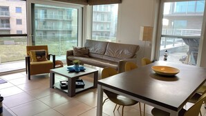 A modern living room with a sofa, armchairs, a coffee table, and a dining table. Large windows offer views of a building and a garden. - Apartment with sunny terrace in Oostduinkerke (OOSTDUINKERKE)