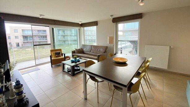 A modern living room with a dining table, sofa, and chairs. Large windows provide a view of the outside. - Apartment with sunny terrace in Oostduinkerke (OOSTDUINKERKE)