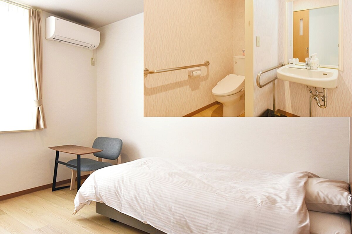 A hotel room with a bed, a chair, a small table, a window with curtains, an air conditioning unit, a toilet, and a sink with a mirror.