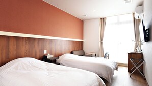 Ein Hotelzimmer mit zwei Betten, einem hölzernen Kopfteil, einem kleinen Tisch, einem Sessel, einem Schreibtisch mit Computer und einem an der Wand befestigten Fernseher.