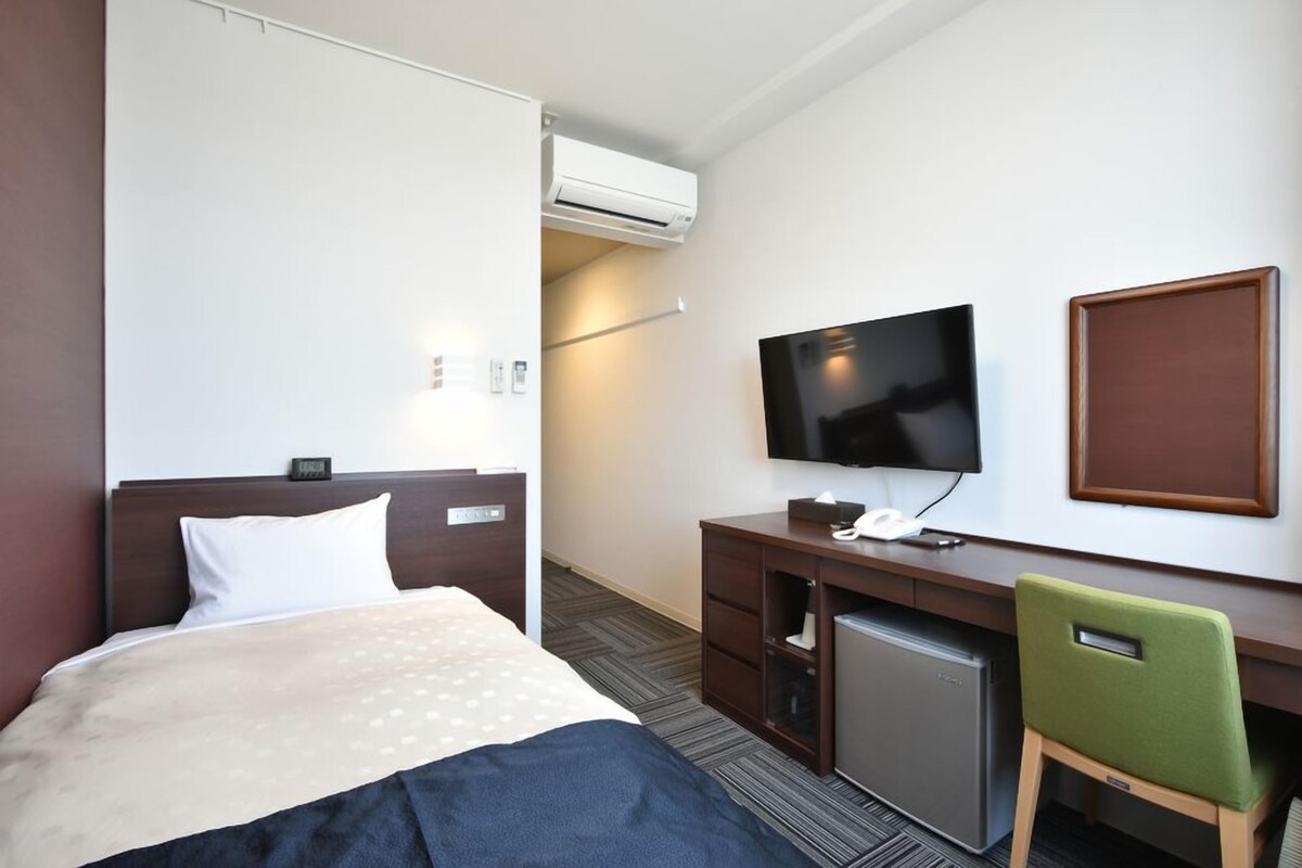 A hotel room with a bed, a desk with a television, a small refrigerator, and a green chair.
