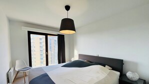 Moderni hotellihuone, jossa on suuri sänky, yöpöytä, tuoli ja ikkunasta avautuva näkymä korkeaan kerrostaloon.