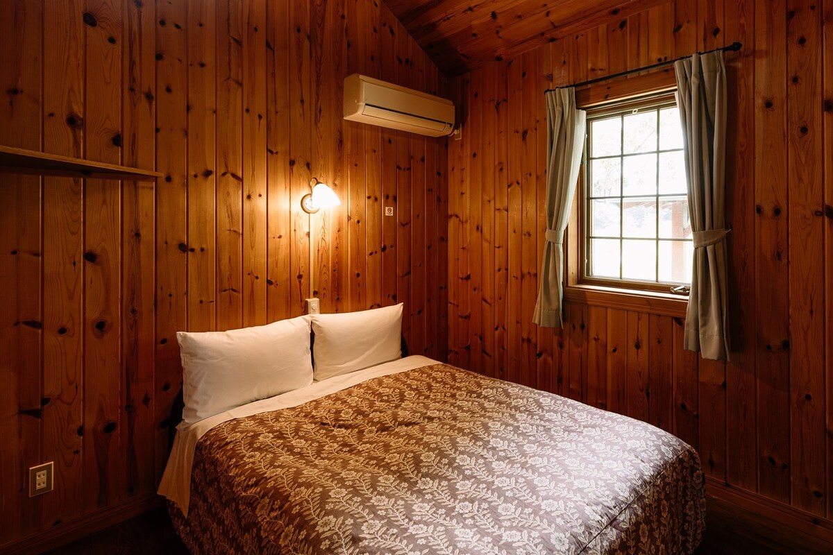 A wooden-paneled room with a bed, a window with curtains, and an air conditioning unit.