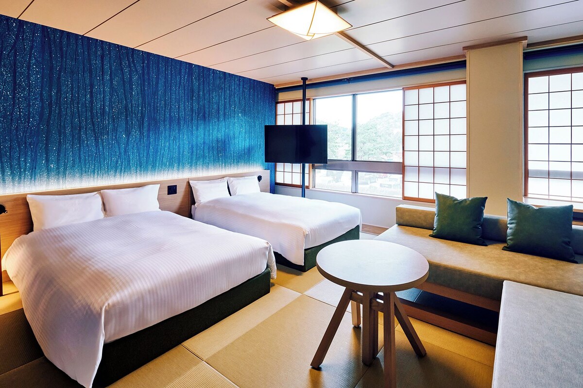 A hotel room with two beds, a sofa, a small table, and large windows.