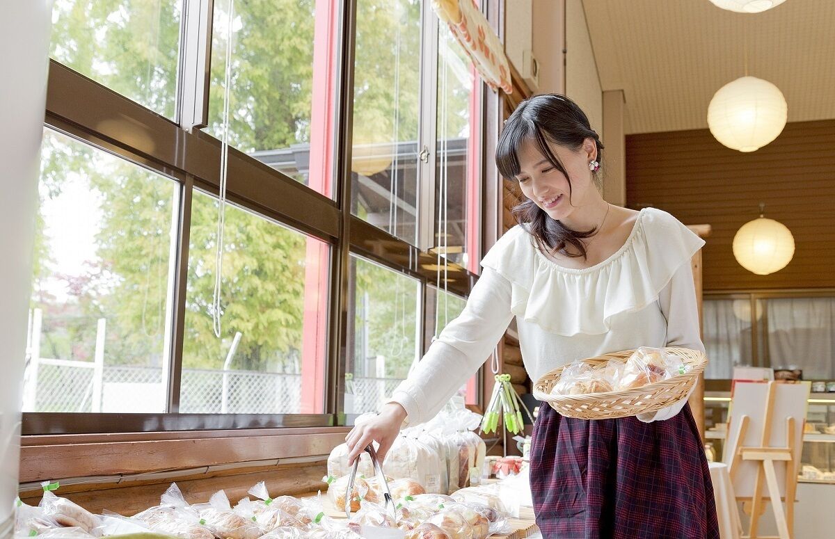 A woman in a white blouse and plaid skirt arranging bread in a basket in a room with large windows and wooden beams.