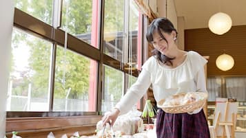 A woman in a white blouse and plaid skirt arranging bread in a basket in a room with large windows and wooden beams.