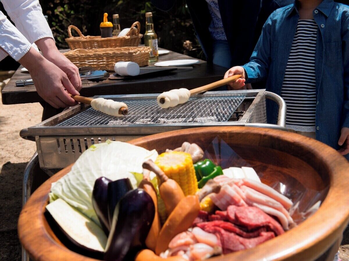 A grill with various meats and vegetables, a basket with condiments, and a person grilling.