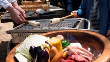 A grill with various meats and vegetables, a basket with condiments, and a person grilling.