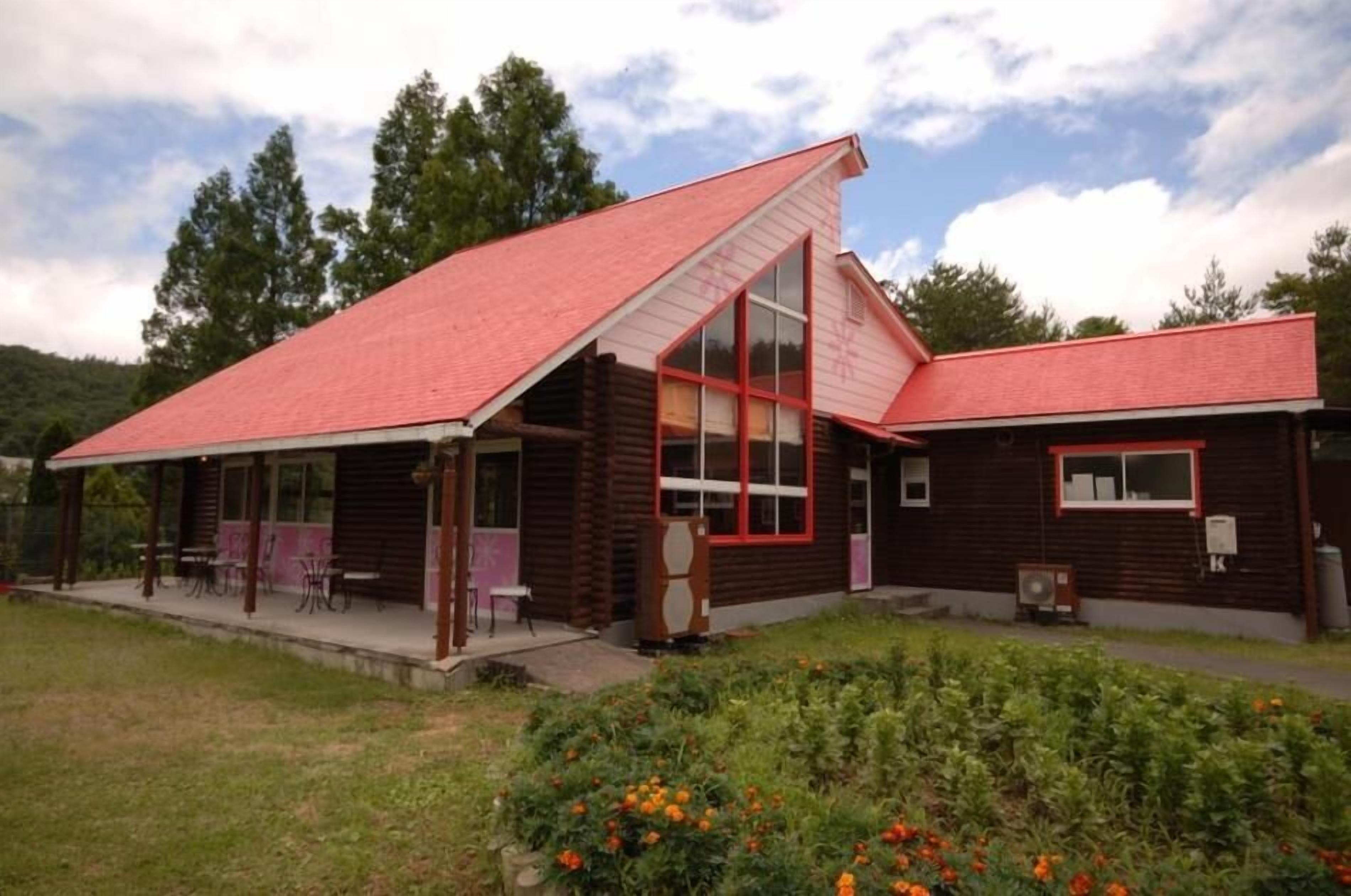A wooden cabin with a red roof, large windows, and a porch with chairs.