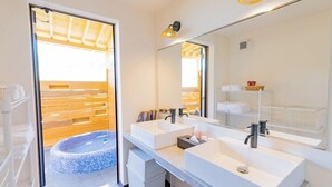 Et moderne badeværelse med to håndvaske, et stort spejl, og en sauna, der kan ses igennem en åben dør.