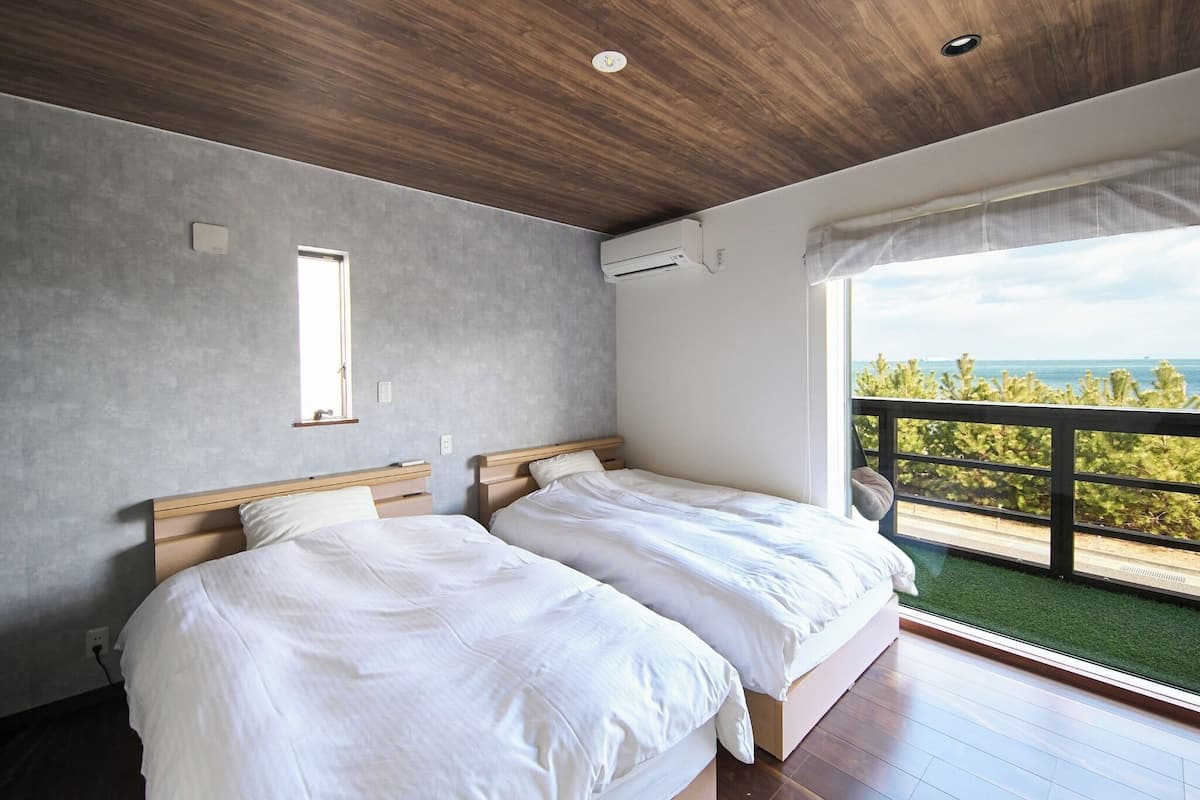 A bedroom with two beds, a wooden ceiling, a grey wall, and a balcony with a view of the sea.