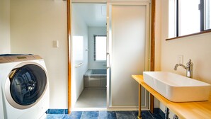 Een wasruimte met een wasmachine, een kleine badkamer met een wastafel en raam, en een houten bank.
