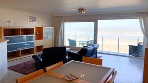 A room with a large window, a ceiling fan, a TV, and a seating area with a view of the beach. - Sea-view apartment on Zeedijk (NIEUWPOORT)