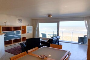 A room with a large window, a ceiling fan, a TV, and a seating area with a view of the beach. - Sea-view apartment on Zeedijk (NIEUWPOORT)