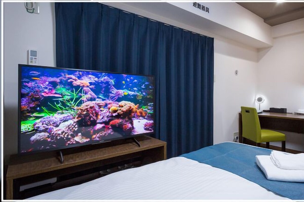 A hotel room with a bed, a desk, a chair, a television displaying an underwater scene, and a curtain.