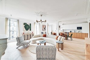 Un salon moderne avec un canapé d’angle, des fauteuils, une table basse et un coin repas, la cuisine étant visible à l’arrière-plan.