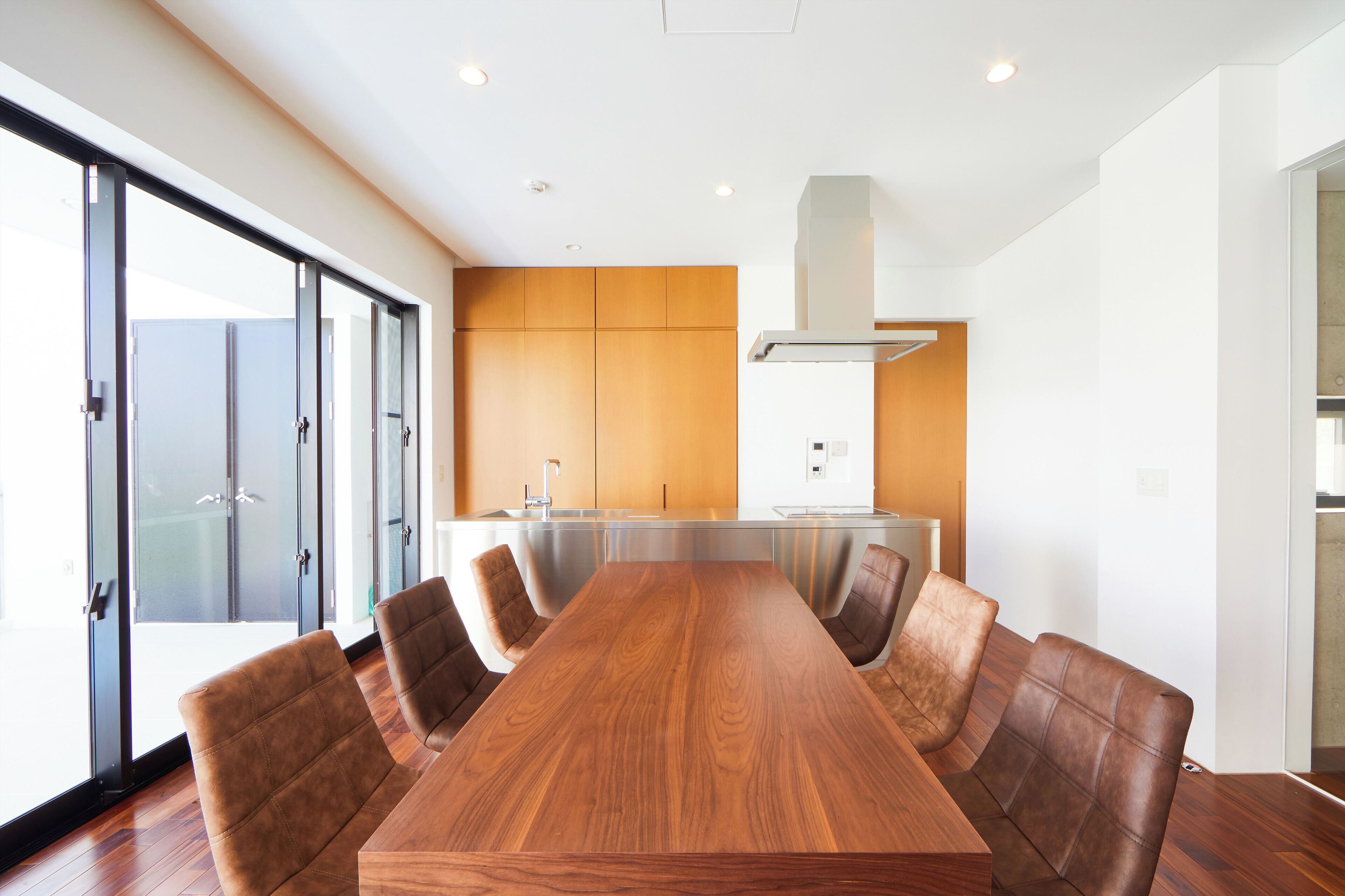 A modern conference room with a wooden table and leather chairs, large windows, and a kitchen area in the background.