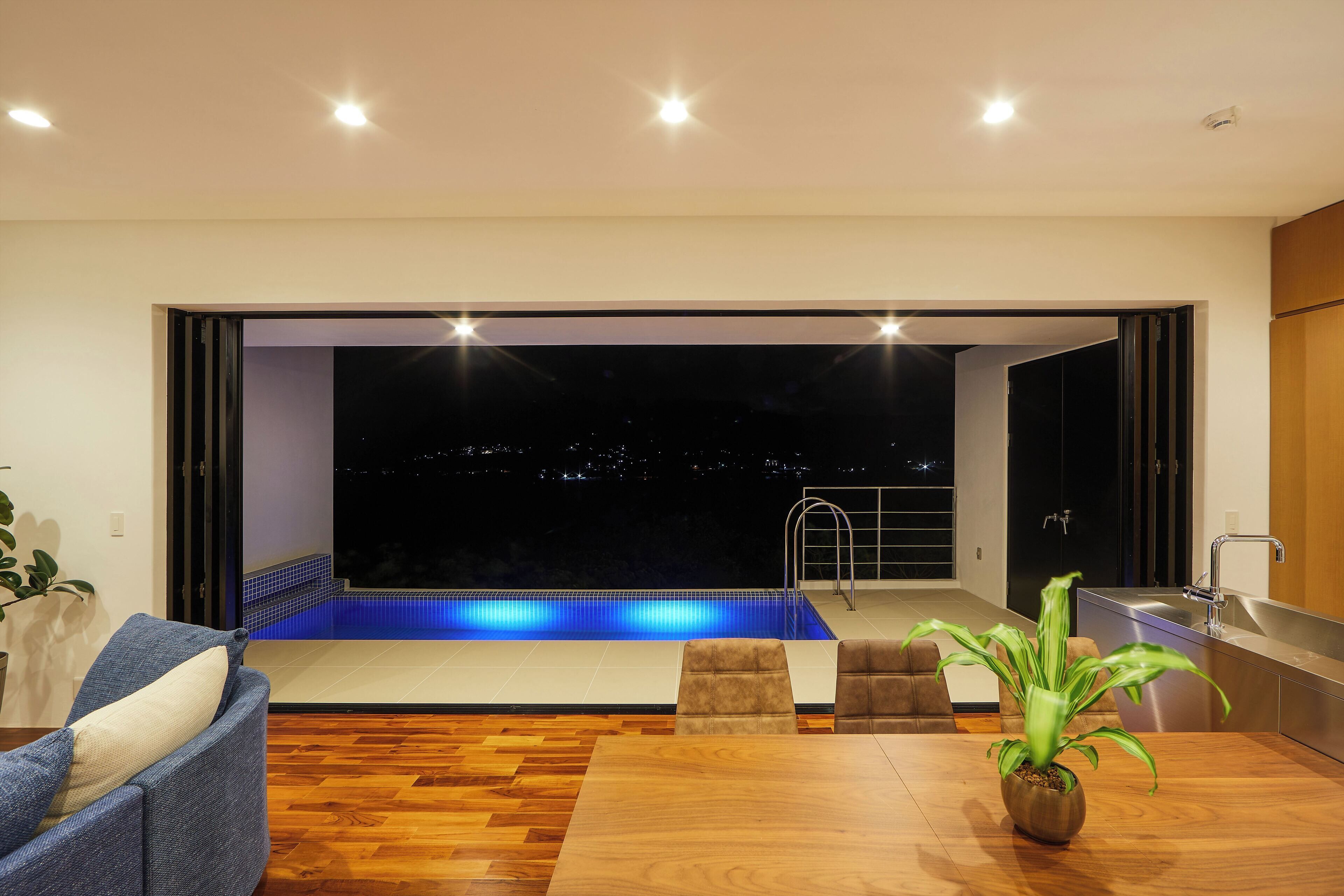 A modern living room with a wooden floor, a blue-lit pool, and a city view at night.