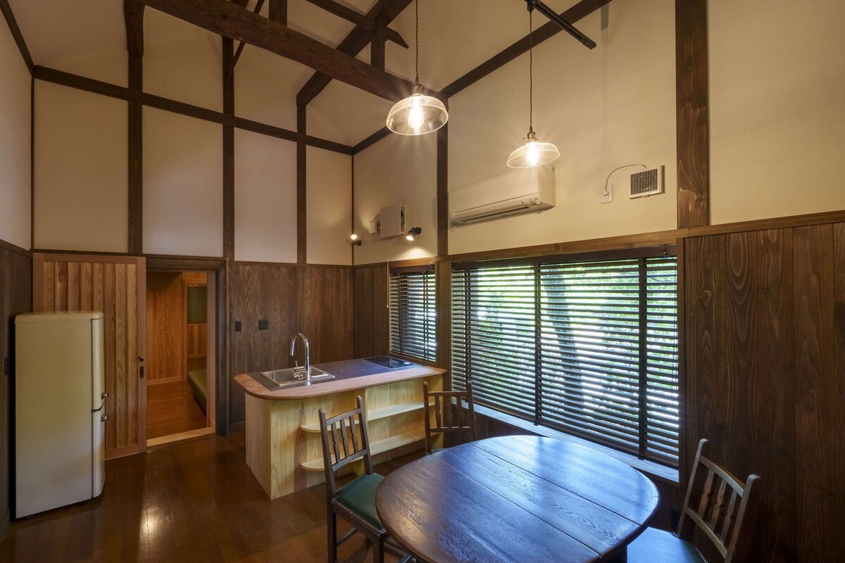 A compact kitchen with a wooden table and chairs, a refrigerator, and a window with blinds.