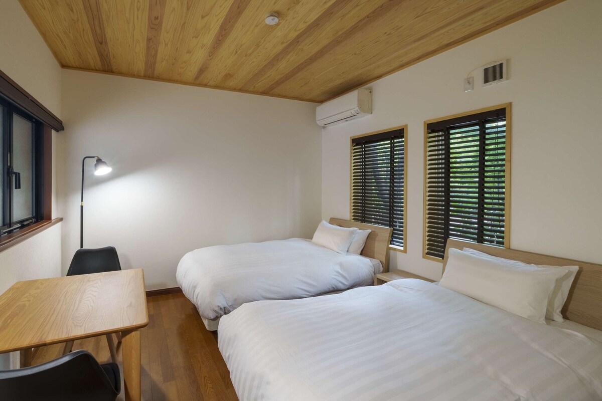A bedroom with two beds, a desk, a chair, a floor lamp, and windows with blinds.