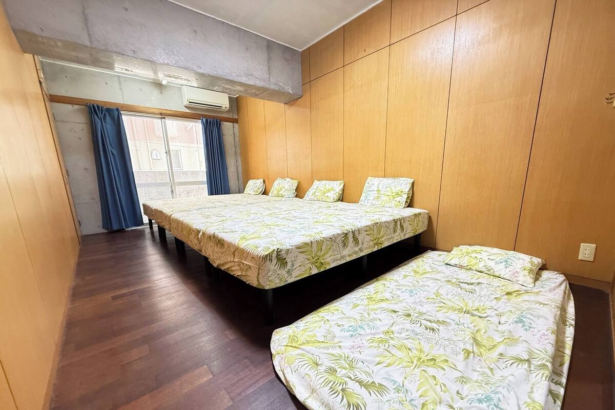 A room with two beds, wooden walls, a window with curtains, and an air conditioning unit.