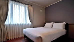 Siististi pedattu sänky hotellihuoneessa, jossa on suuri ikkuna, verhot ja puulattia.