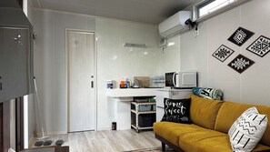 A compact kitchen with a microwave, a coffee machine, and a small refrigerator. There is a white countertop and a yellow sofa with patterned cushions. - Tabijiman (Nago)
