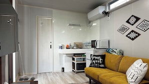 A compact kitchen with a microwave, a coffee machine, and a small refrigerator. There is a white countertop and a yellow sofa with patterned cushions. - Tabijiman (Nago)