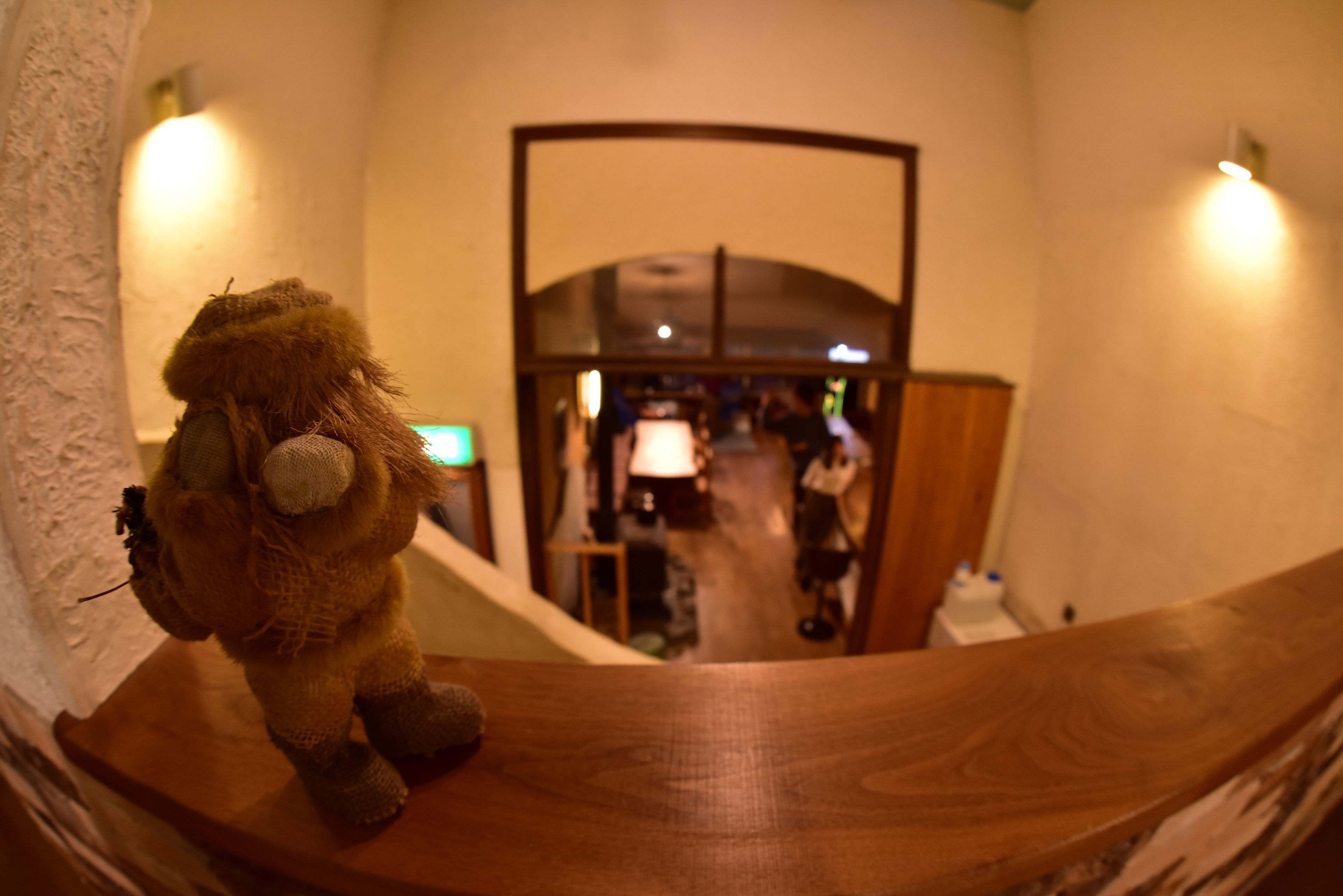 Un peluche sobre el barandal de una escalera de madera, con vistas a un pasillo que tiene una puerta y una persona a lo lejos.