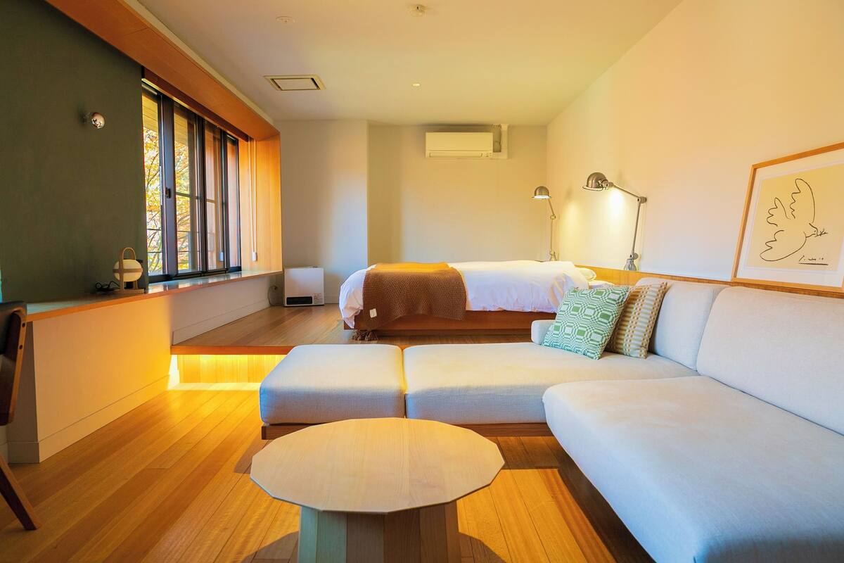 A modern hotel room with a large bed, a sofa, a round table, and a window with blinds.